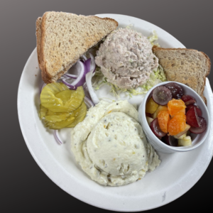 picture of a tuna salad plate which consist of a scoop of tuna, potato salad, fresh fruit, toast, pickles, and red onions.